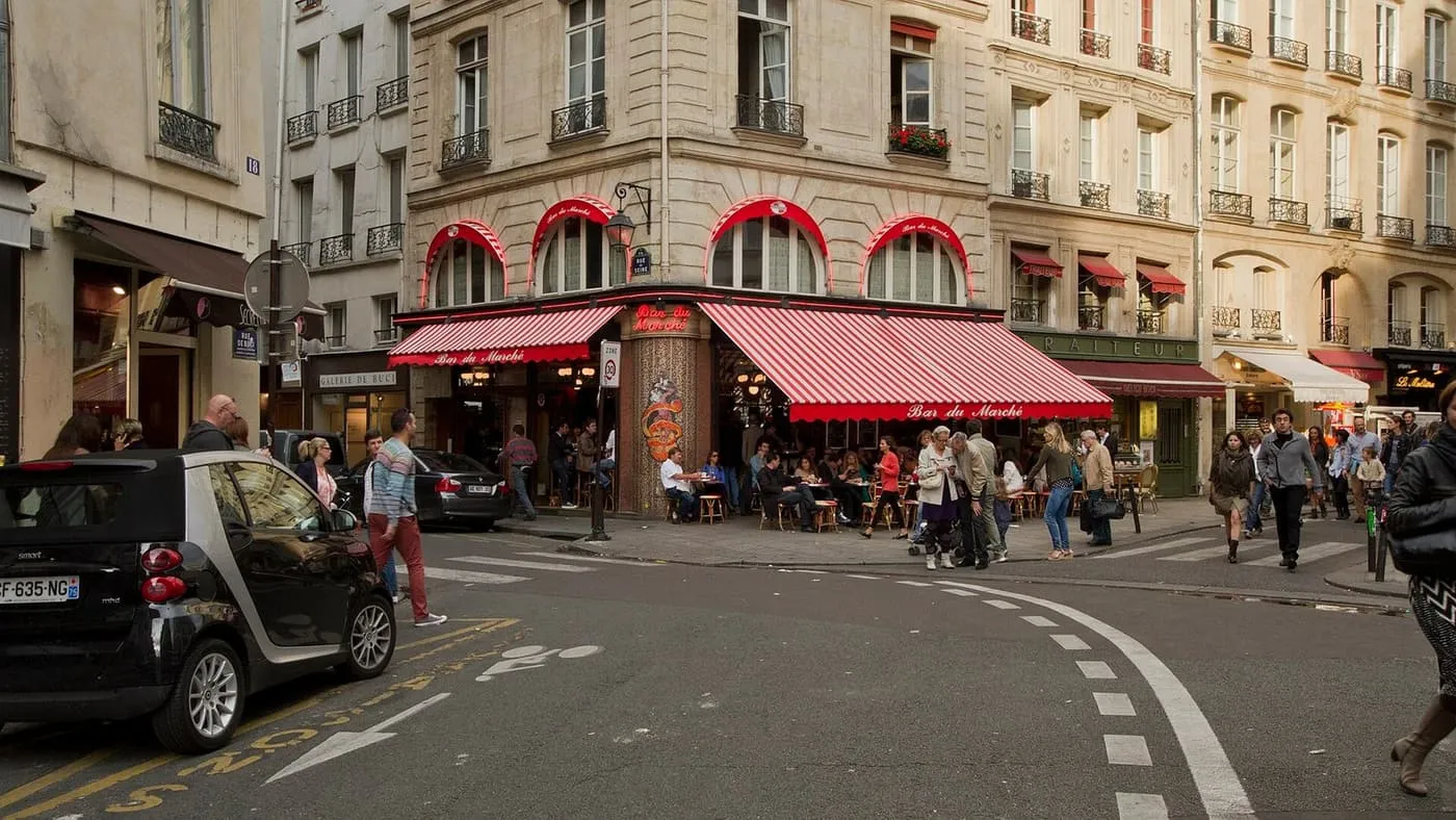 A corner café with red-and-white striped awnings and terrace tables where Rue de Buci meets Rue de Seine, with people crossing the street