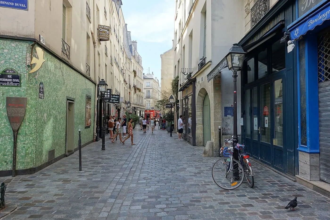 A cobbled street in Le Marais, with a green mosaic wall on the left, a blue boulangerie shopfront on the right and people walking towards a corner ahead