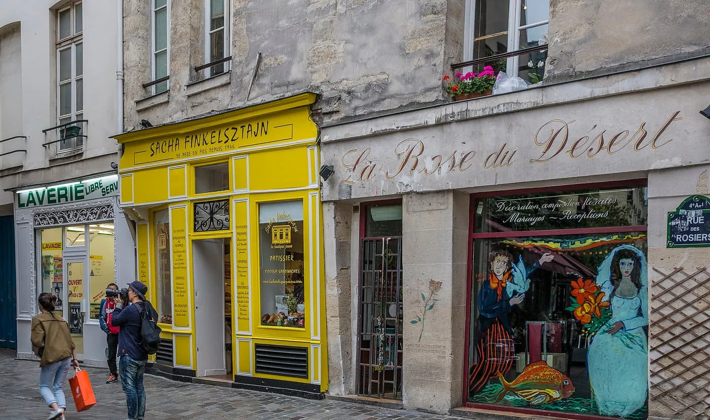 A cobbled stretch of rue des Rosiers, with a yellow shopfront, a painted florist's window and a street sign for the rue des Rosiers