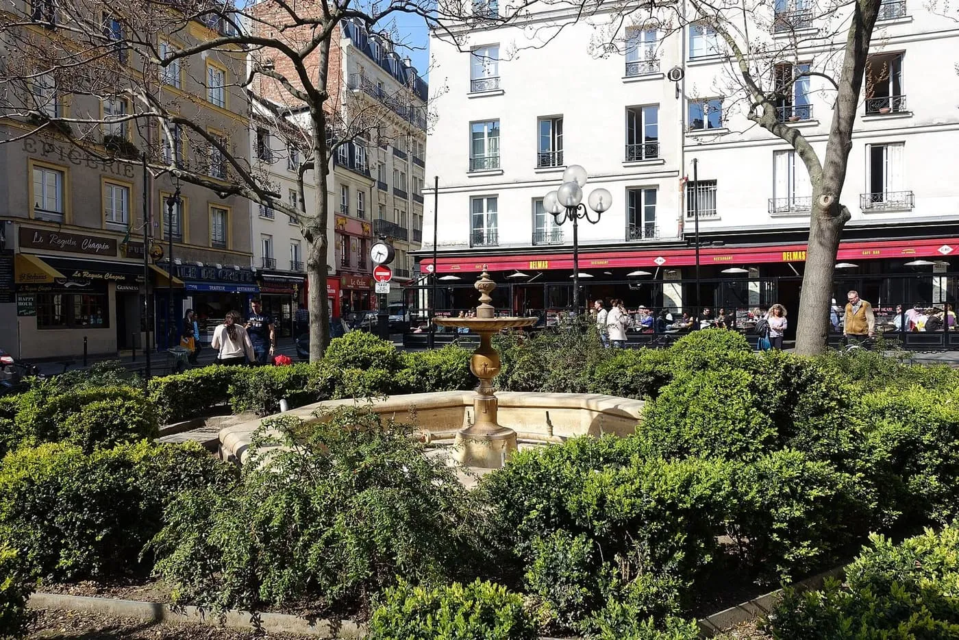 A stone fountain in a small planted square, with café terraces and shopfronts behind, at the top of Rue Mouffetard