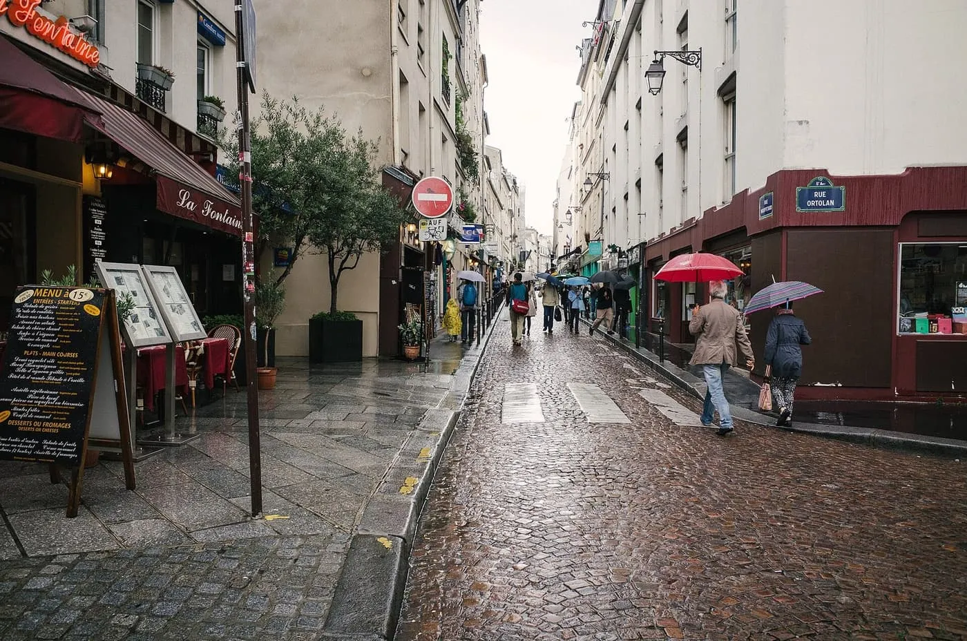 Rue Mouffetard on a wet day, with restaurant terraces on the left, cobbles ahead and people walking under umbrellas