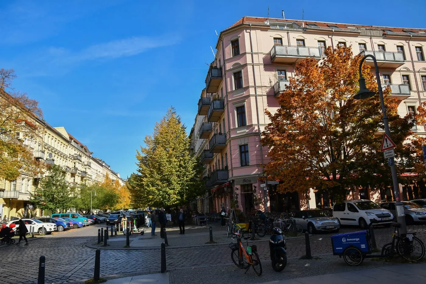 A corner of Rykestraße in Prenzlauer Berg with a pink Gründerzeit apartment block, autumn trees and cobbled crossing