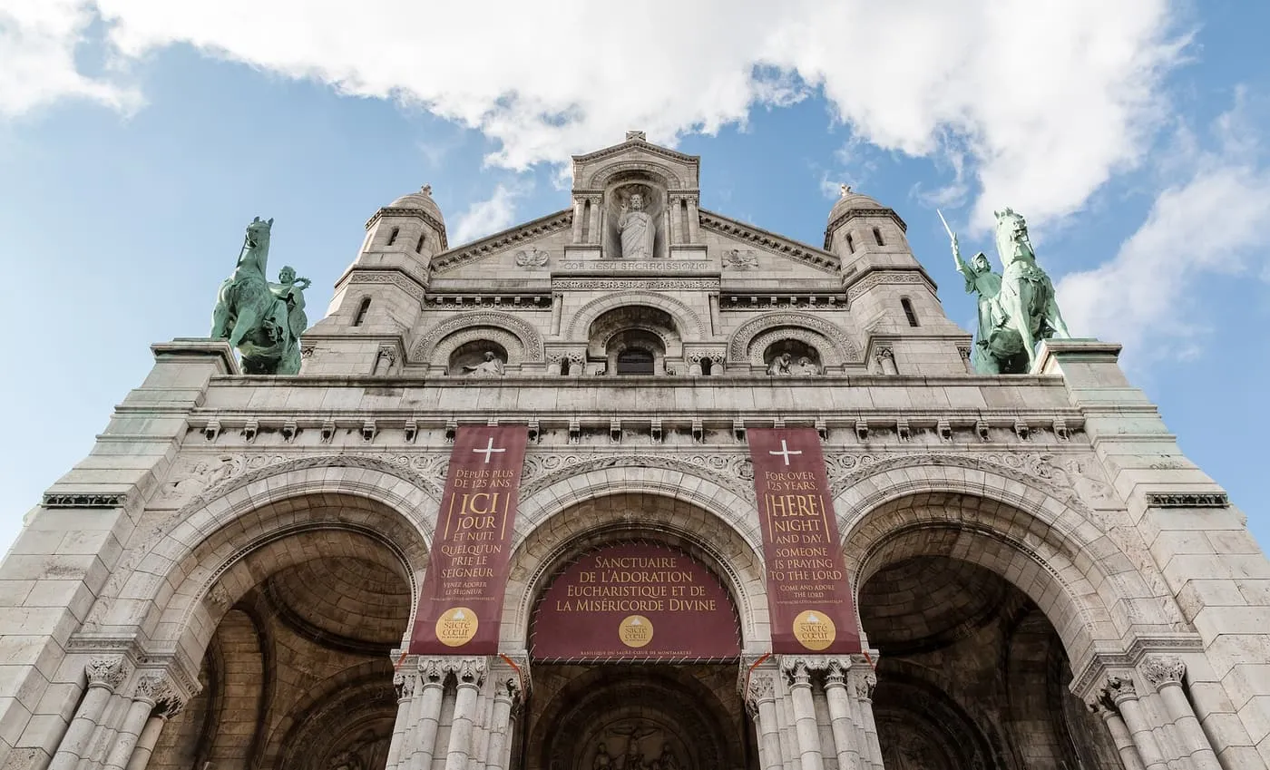 The front of the Sacré-Cœur seen from below, three arched doorways under a gallery with two green equestrian statues, and red banners hung across the entrance