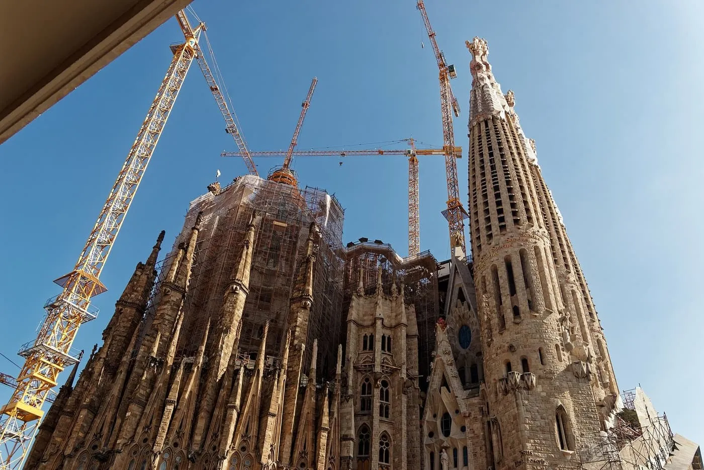 Looking up at the towers of the Sagrada Família from Carrer de Provença, with building cranes beside them against a blue sky