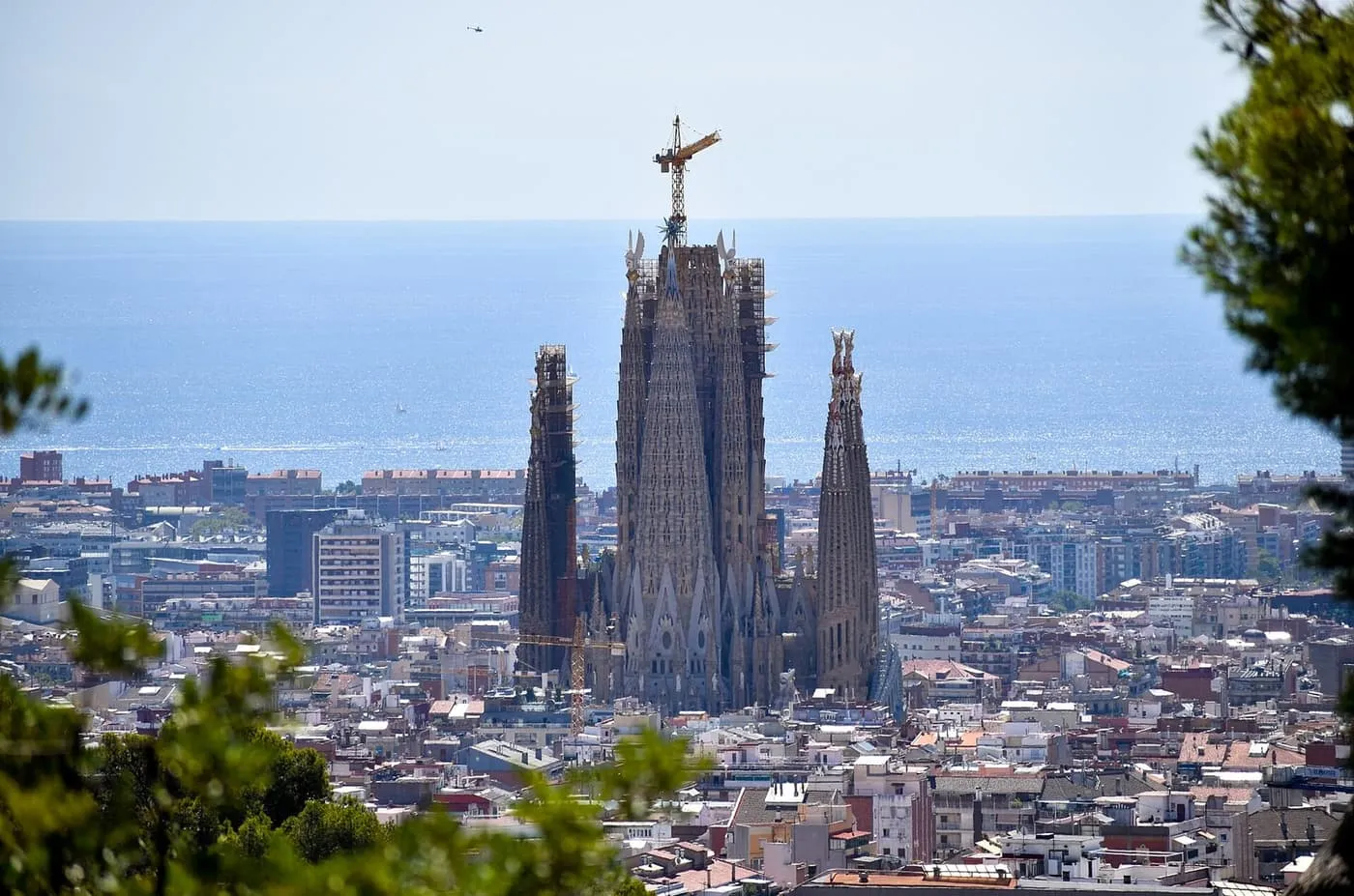 The spires of the Sagrada Família, one topped by a crane, rising above the city with the sea behind