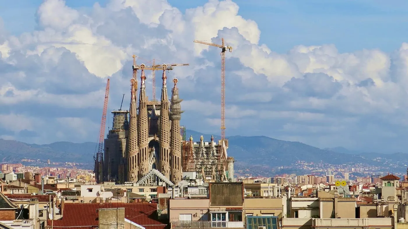 The towers of the Sagrada Família with construction cranes rising over the rooftops of the Eixample, with hills and cumulus cloud behind