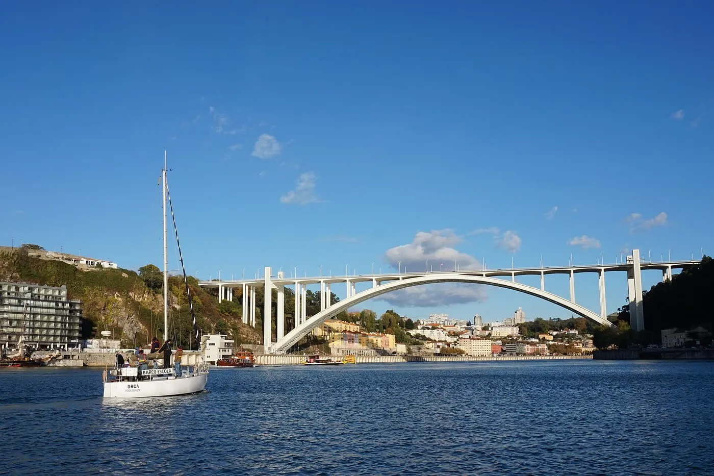 A white sailing boat on the Douro in front of the Arrábida bridge, whose single concrete arch spans the river under a blue sky