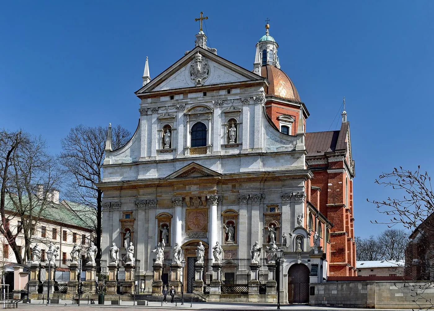 The white stone Baroque facade of the Church of Saints Peter and Paul behind a railing lined with statues of the apostles