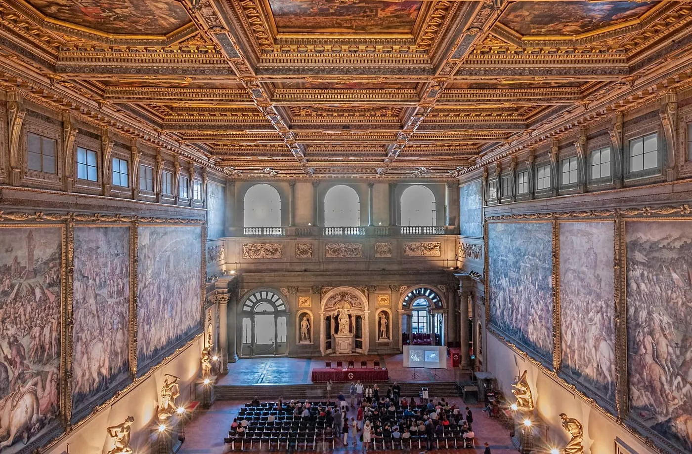 The Salone dei Cinquecento seen from its gallery, with a gilded coffered ceiling, battle frescoes on both long walls and rows of chairs on the floor