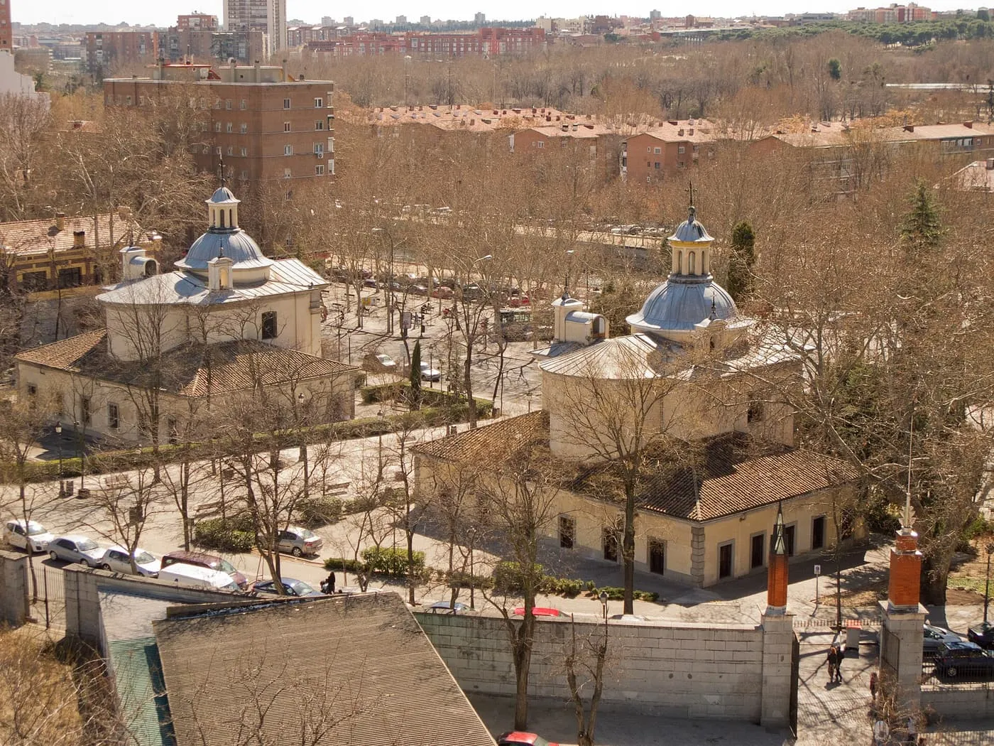 The two small domed chapels of San Antonio de la Florida standing either side of a street among bare trees, seen from above