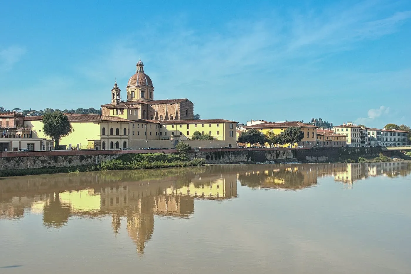The dome and bell tower of San Frediano in Cestello above a row of yellow buildings on the Arno, reflected in the river