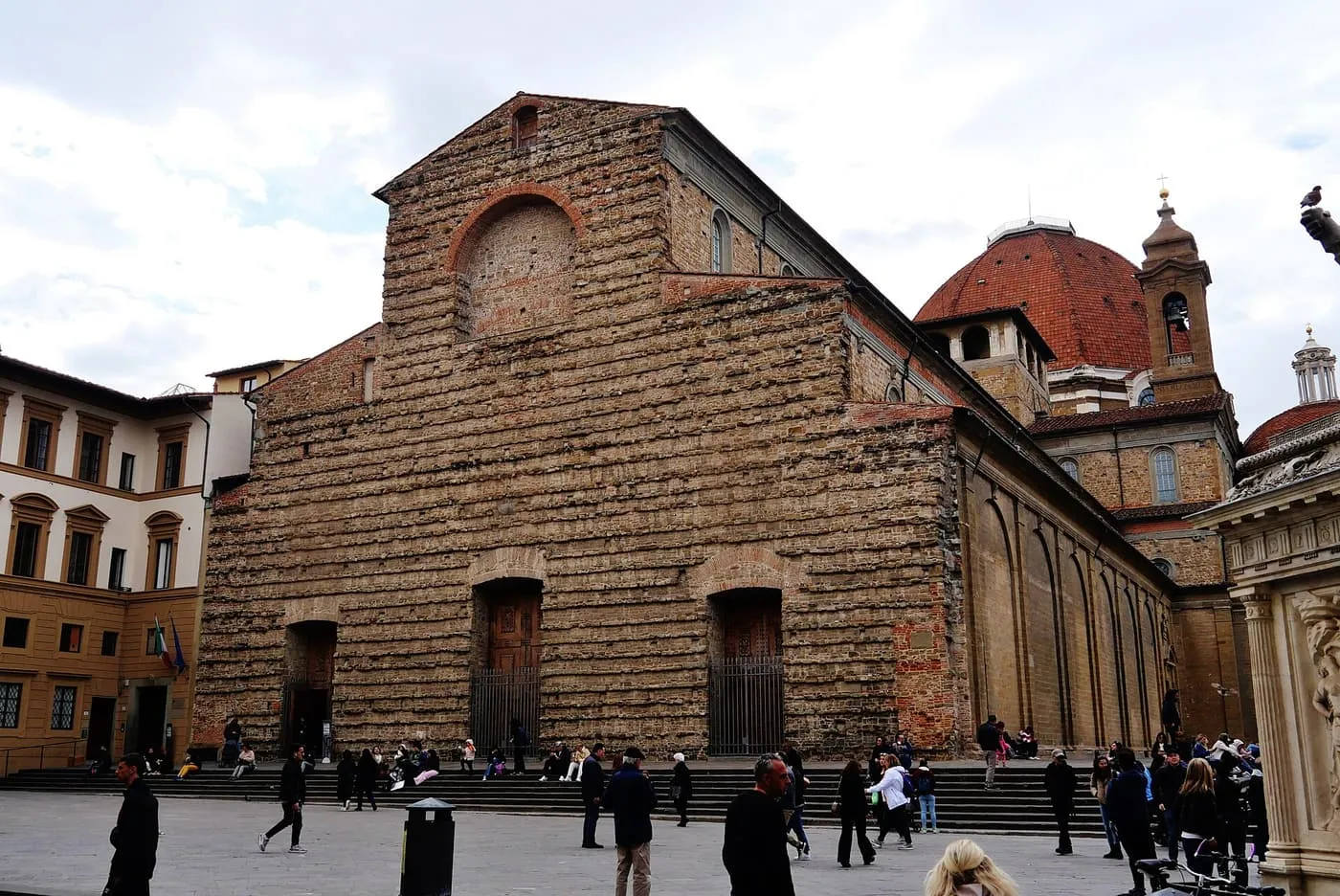 The unfinished rough-stone front of San Lorenzo with a dome rising behind it and visitors on the steps
