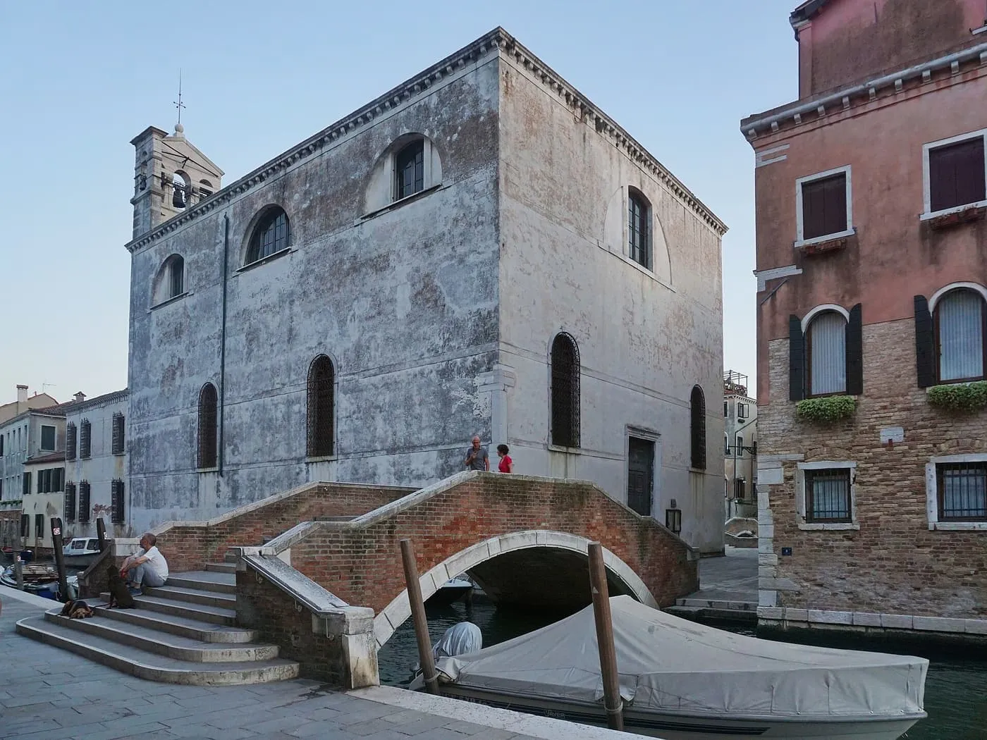 A plain stone building with a small bell-gable beside a stepped brick bridge over a canal