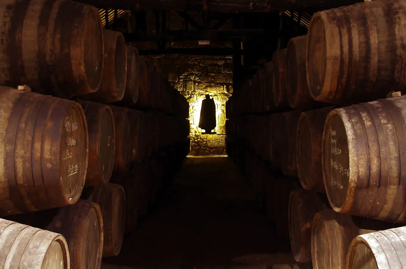 Rows of old port casks in a dark stone cellar, with a figure in a black cape and hat silhouetted in a doorway at the end