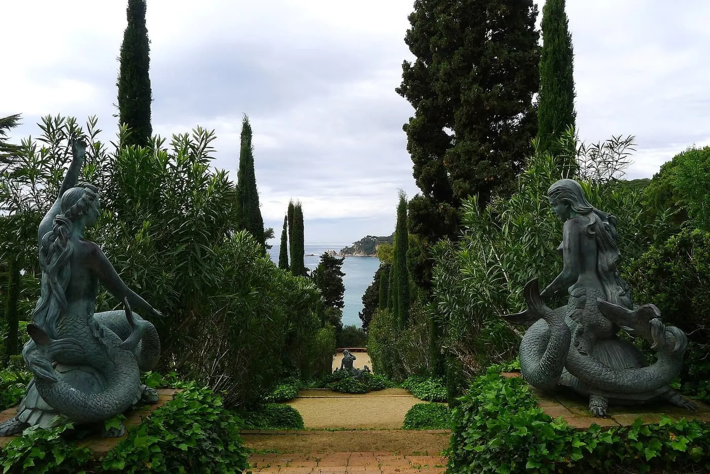 Two bronze mermaid statues on either side of a garden path between cypress trees, with the sea in the gap ahead