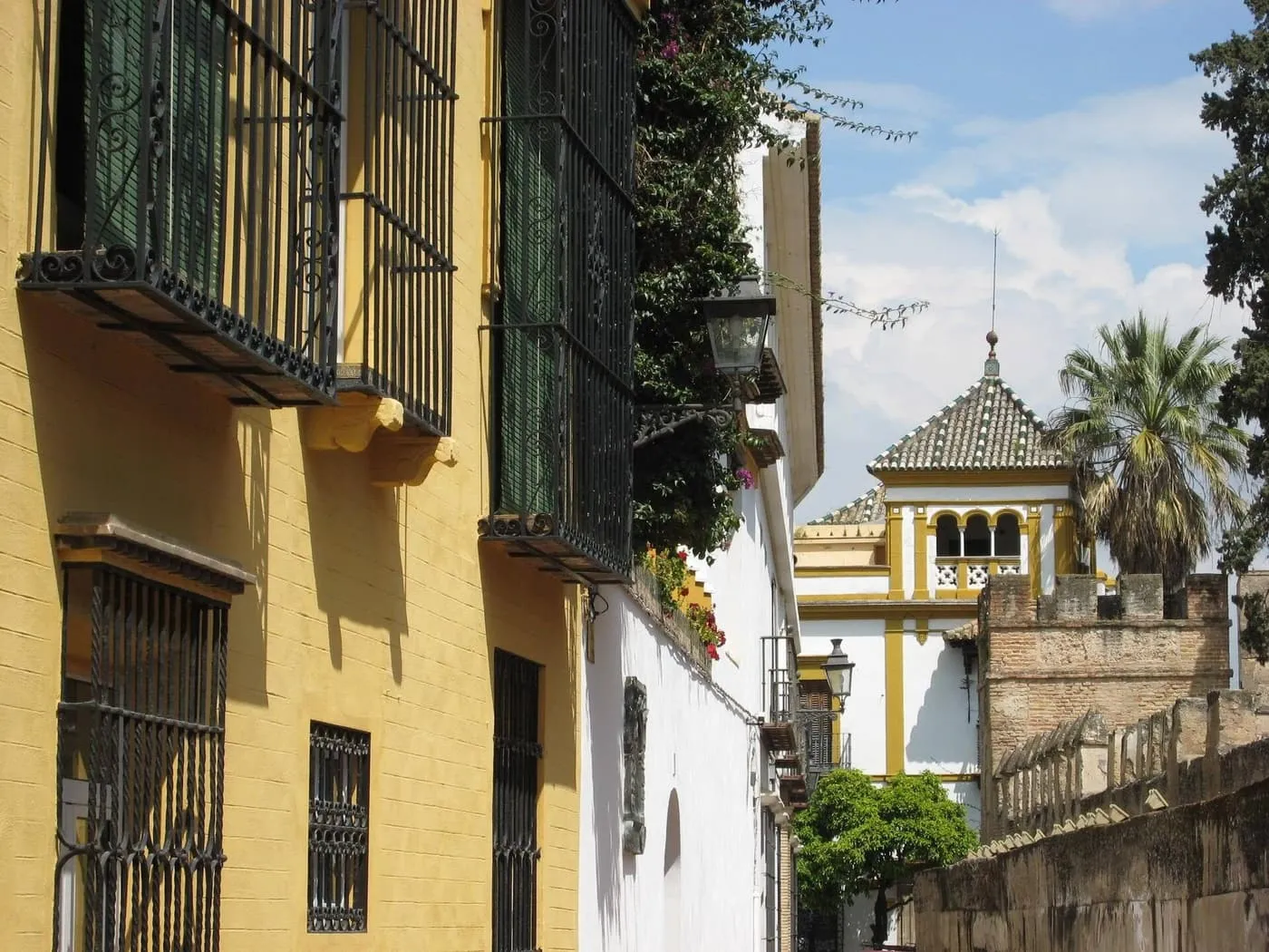 A narrow lane in Santa Cruz between an ochre house with green-shuttered balconies and a white wall, with a crenellated brick wall and a tiled turret at the far end