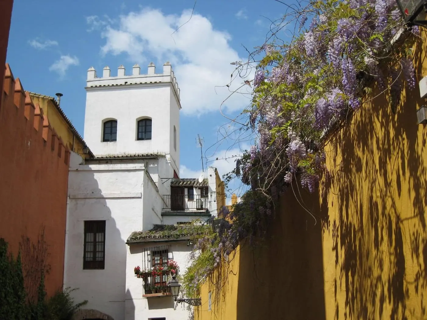 A narrow street in the Santa Cruz quarter with a red crenellated wall on the left, a whitewashed tower and wisteria hanging over a yellow wall