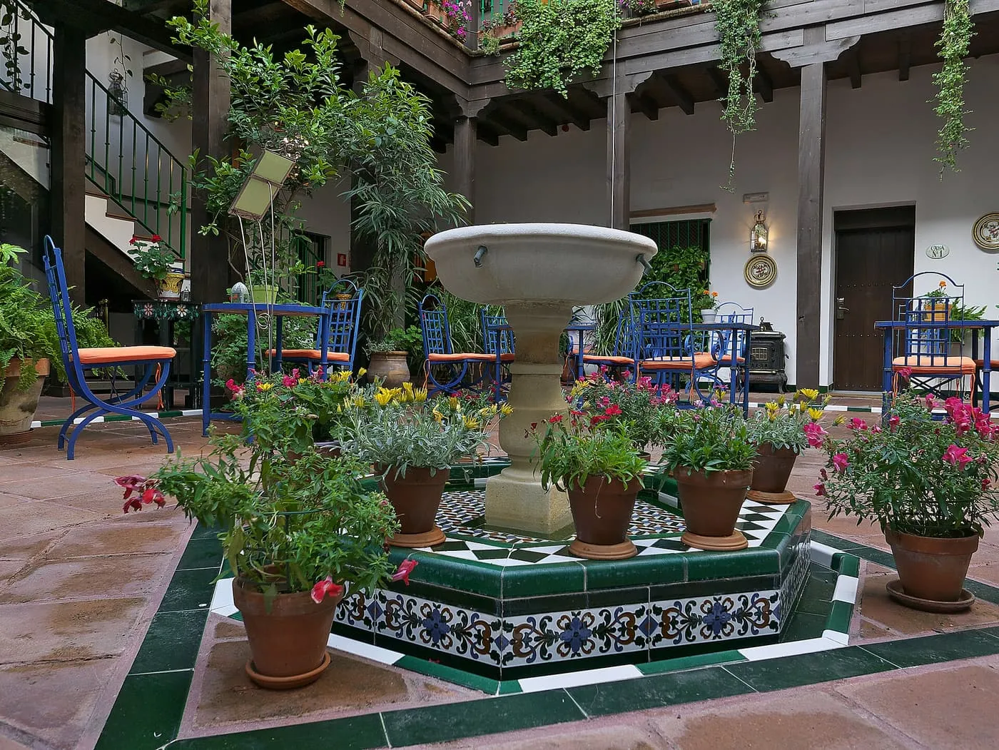 A courtyard patio with a tiled fountain ringed by flowerpots, blue metal chairs and a wooden gallery