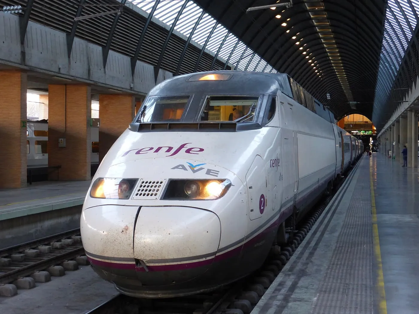 A white Renfe AVE high-speed train at a platform of Seville Santa Justa station, under the curved roof