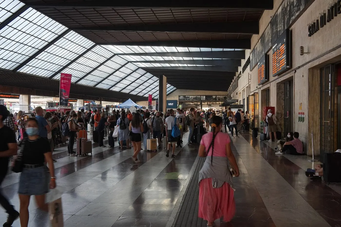 Travellers with luggage crossing the glass-roofed hall of Santa Maria Novella station, with departure boards on the right