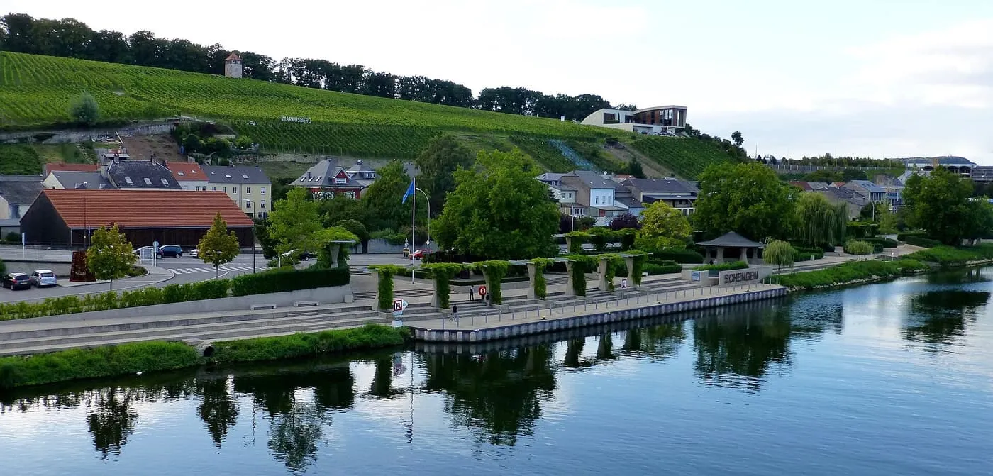 The Moselle waterfront at Schengen, Luxembourg, with a pergola promenade on the quay, the village name on the wall and vineyards on the hill behind