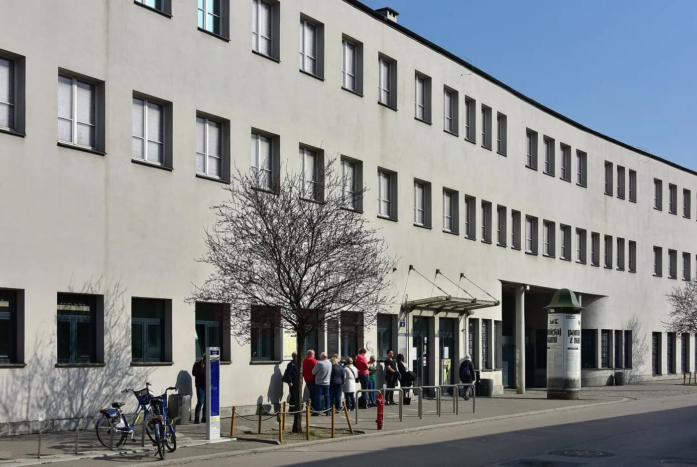 The curved white front of the former administrative building of Schindler's enamel factory on Lipowa Street, with a few visitors waiting at the entrance