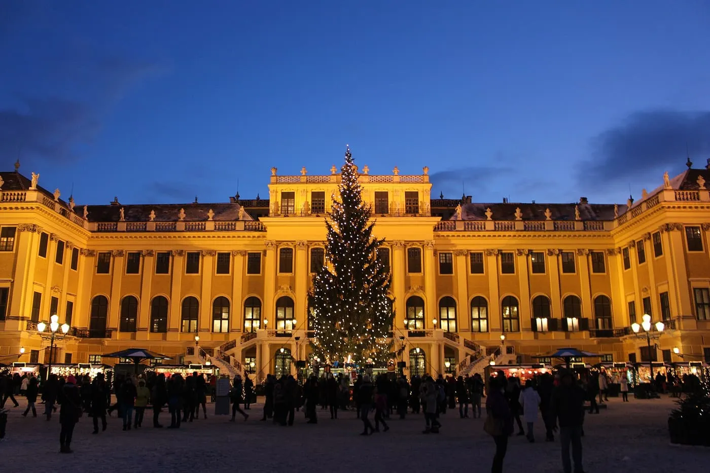 The yellow front of Schönbrunn Palace at dusk with a lit Christmas tree in the courtyard and market stalls along its foot