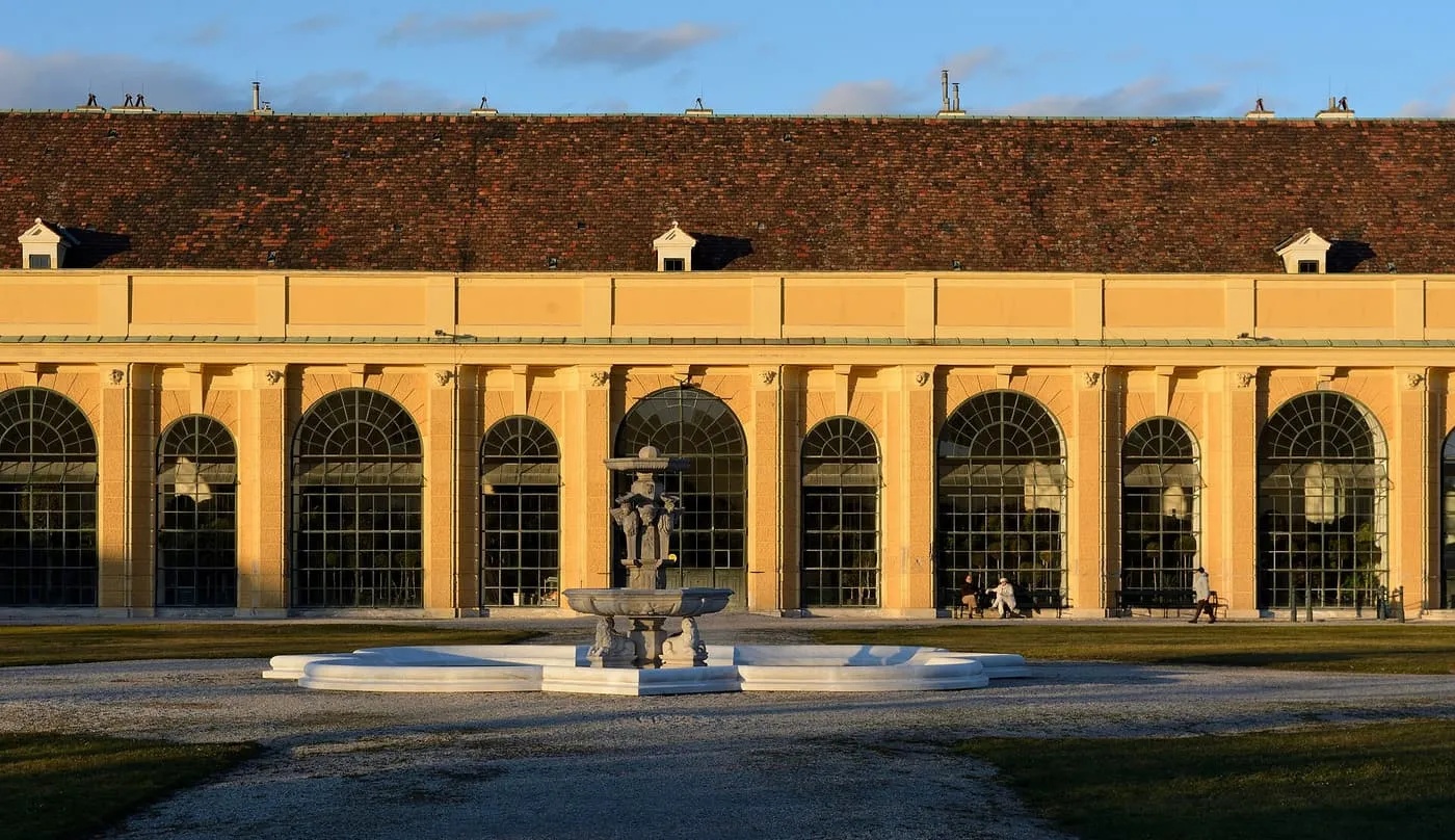 The long yellow façade of the Orangery at Schönbrunn with tall arched windows and a fountain in front, in low evening light