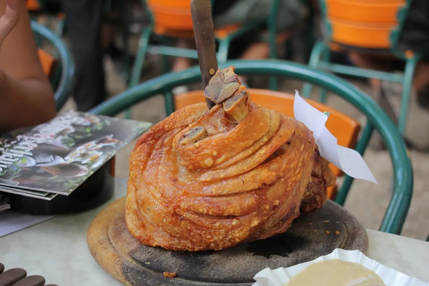 A roast pork knuckle with a crackled skin and a knife stuck in it, on a wooden board on a garden table