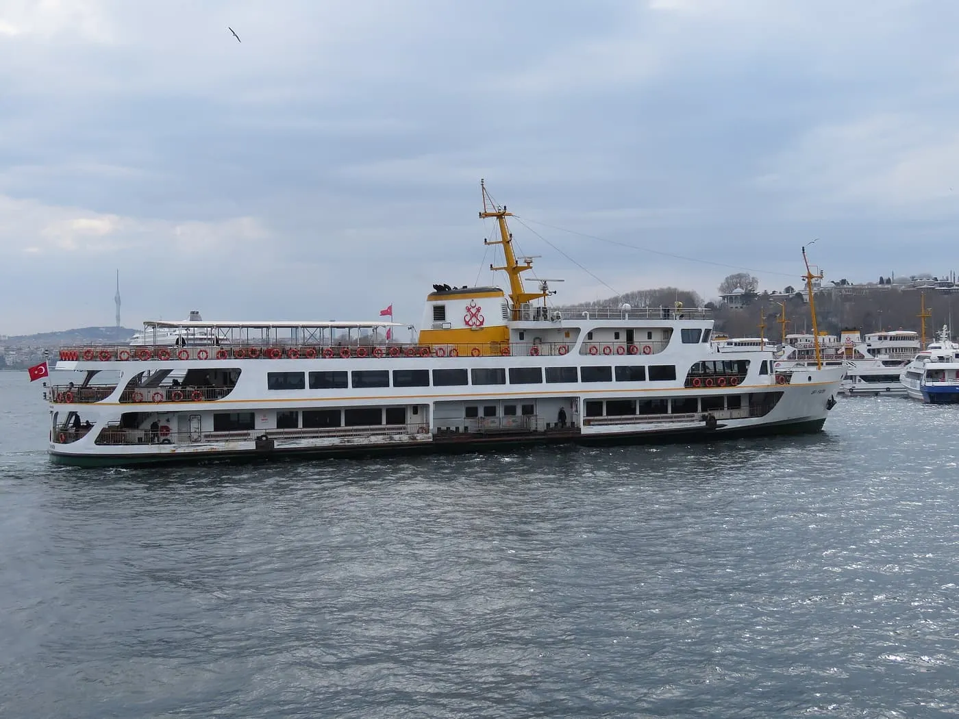 A white Şehir Hatları commuter ferry with a yellow funnel arriving at Eminönü, with other boats moored beyond it