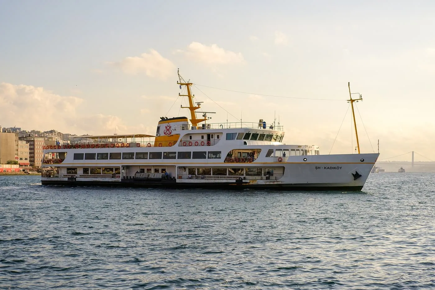A white Şehir Hatları ferry marked Kadıköy crossing the water in warm evening light, with the Bosphorus Bridge faint in the haze behind