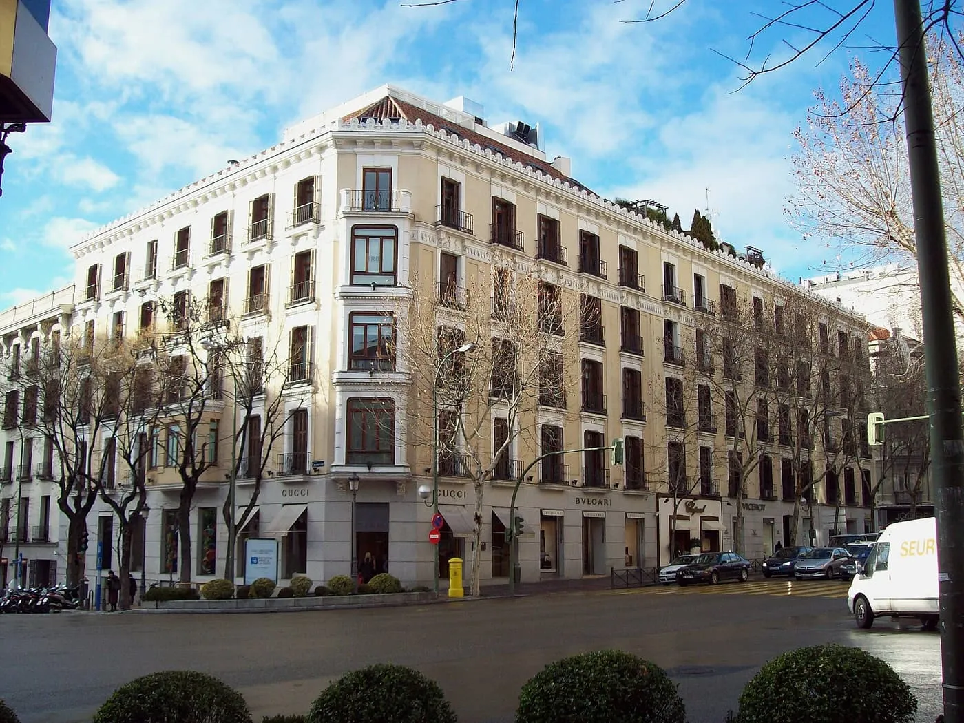 A cream stone corner building on Calle de Serrano with luxury shop fronts at street level and bare plane trees in front