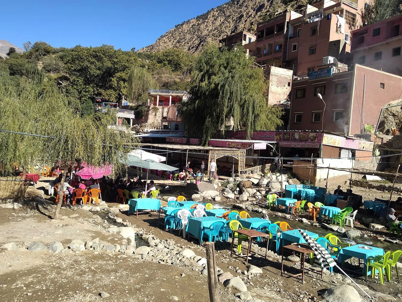 Café tables in turquoise, green and orange plastic beside a stream at Setti Fatma, below pink-walled houses on the hillside