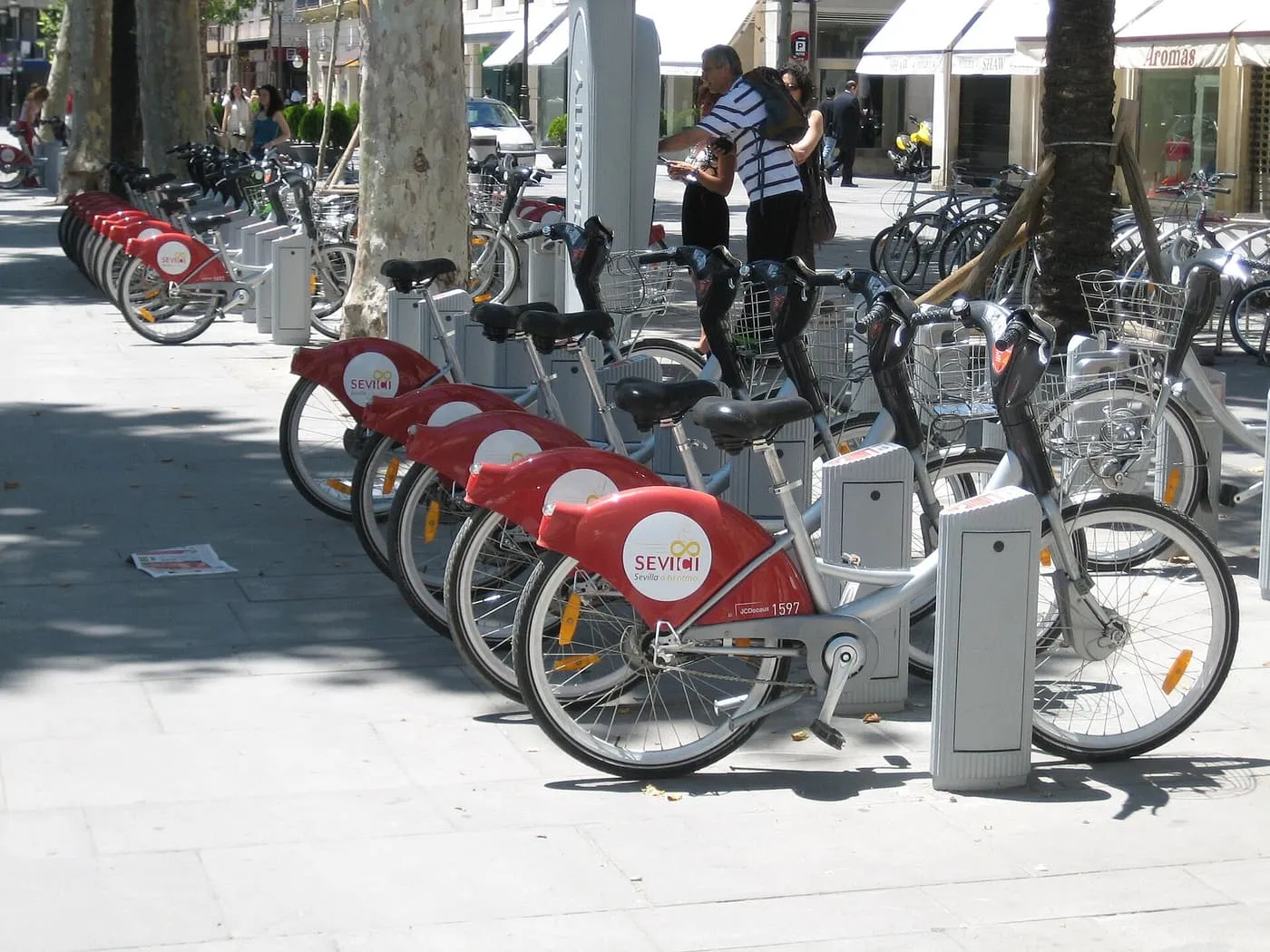 A row of red-and-silver Sevici bikes docked at a station on a shaded Seville pavement