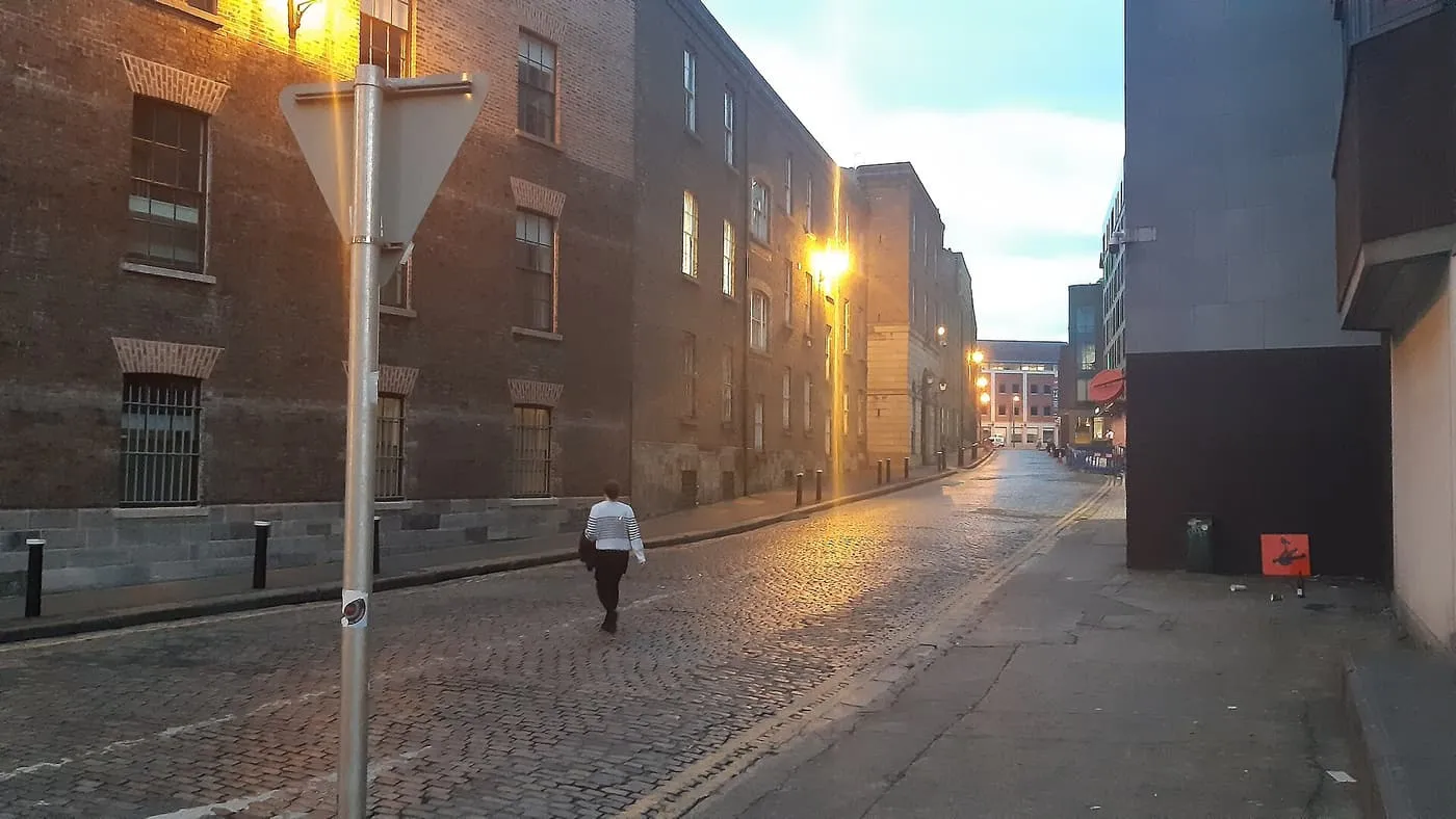 A cobbled street at dusk between brick warehouse-style buildings, lit by warm street lamps, with one pedestrian
