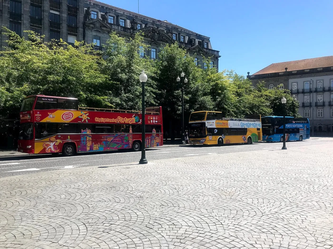 Three open-top sightseeing buses, one red, one yellow and one blue, parked under trees beside a cobbled square