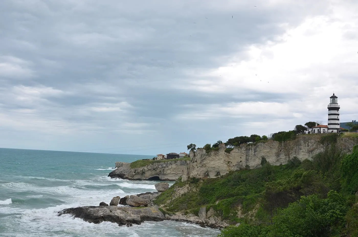 The black-and-white striped Şile lighthouse on a green cliff above a rocky, wave-washed shore