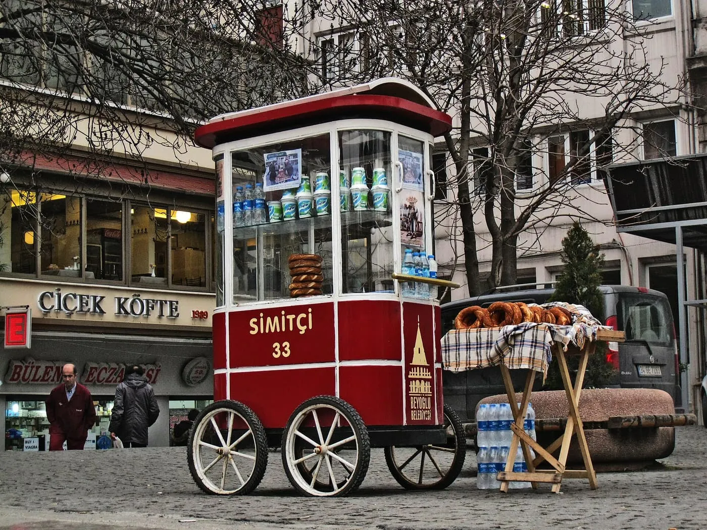 A red glass-fronted simit cart with rings of sesame bread displayed on a table beside it, on a cobbled square