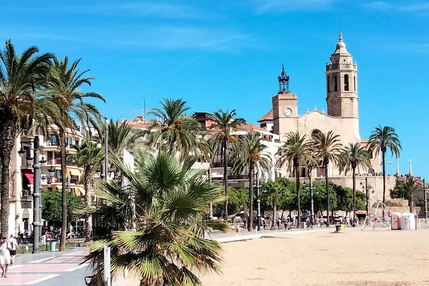The sand of Sitges's seafront beach behind a line of palms, with the church of Sant Bartomeu i Santa Tecla and its bell tower above the promenade