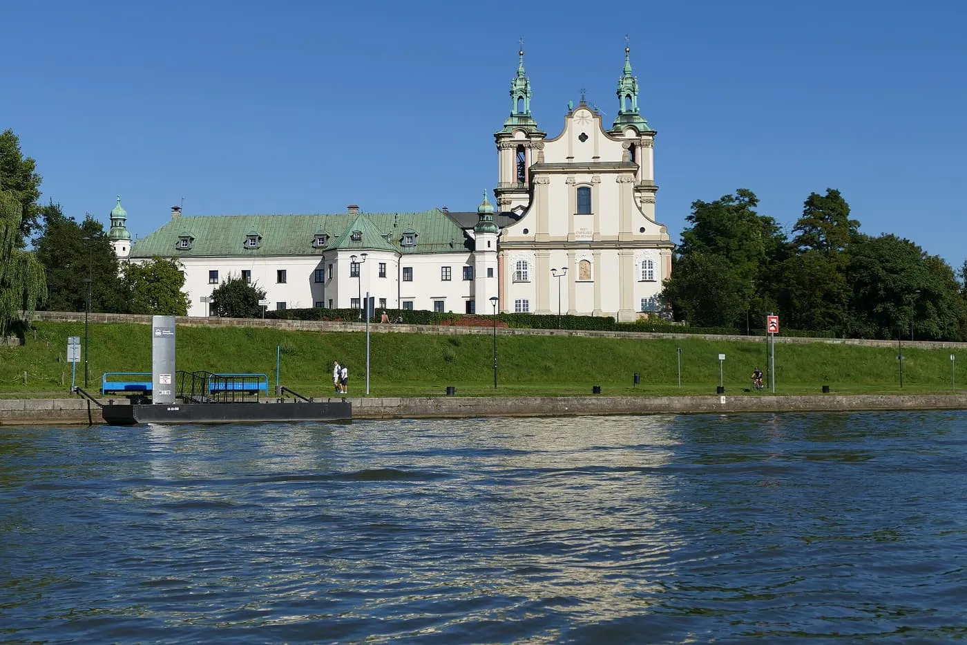 The white baroque church and monastery of Skałka above a grassy embankment, seen across the Vistula from a boat, with a landing stage at the left