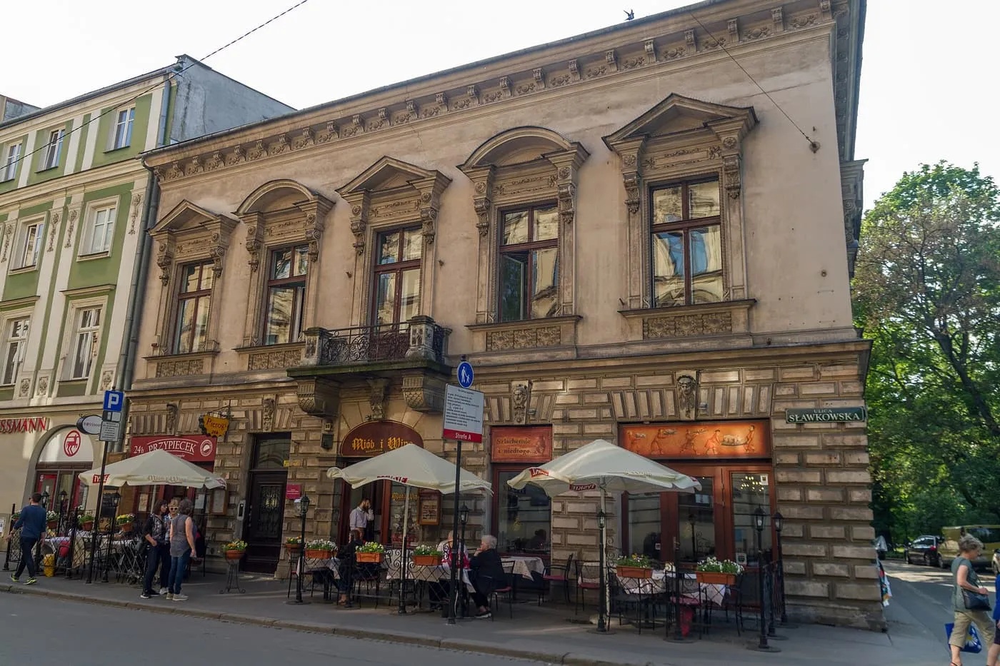 A corner townhouse with rusticated stonework and café terraces under white parasols, beside a street sign reading Sławkowska