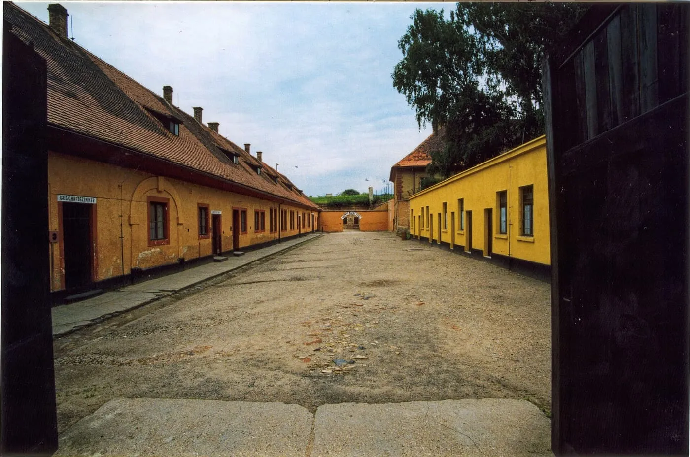 A gravel courtyard in the Small Fortress at Terezín, lined with low yellow and ochre buildings, with an arched gate at the far end