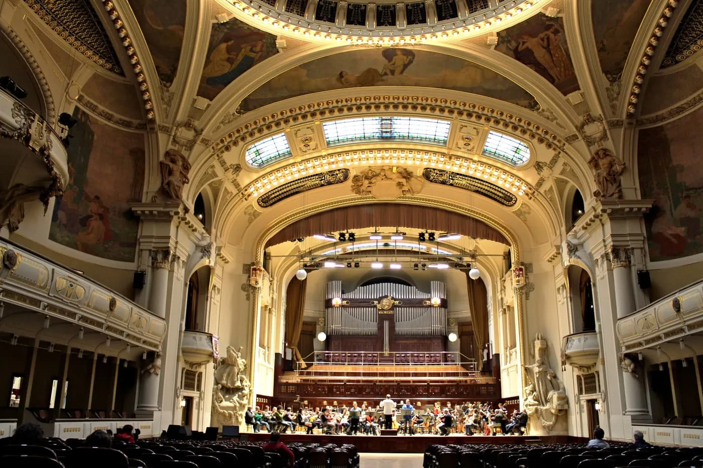 An orchestra rehearsing on the stage of the Smetana Hall in the Municipal House, beneath the organ and a painted Art Nouveau vault