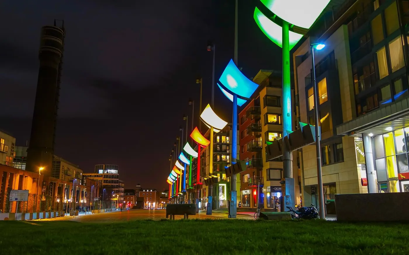 Smithfield Square at dusk with a row of coloured lamp posts, new apartment blocks and a tall brick chimney