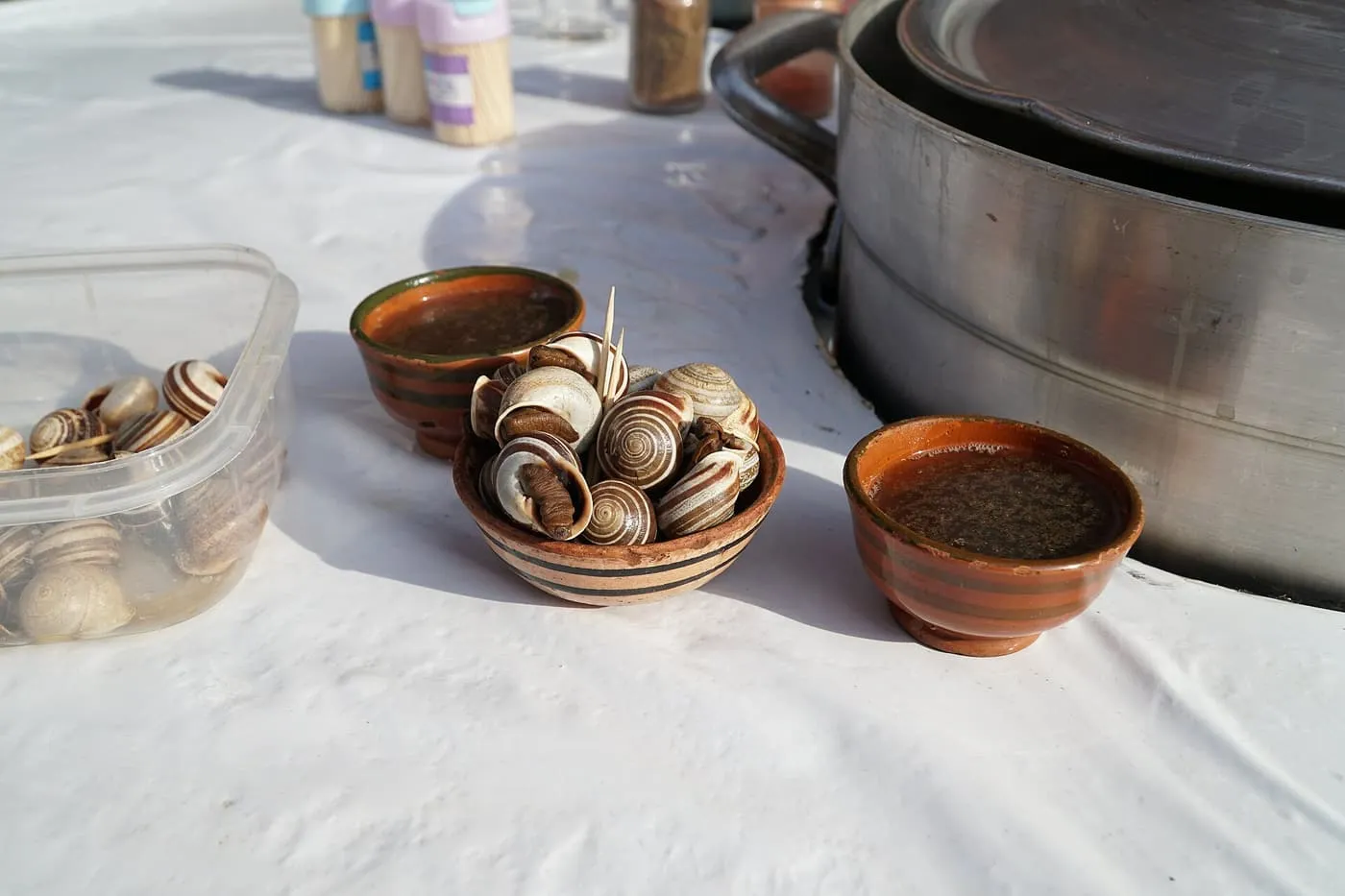 A small bowl of snails in their shells beside two bowls of broth on a white stall counter