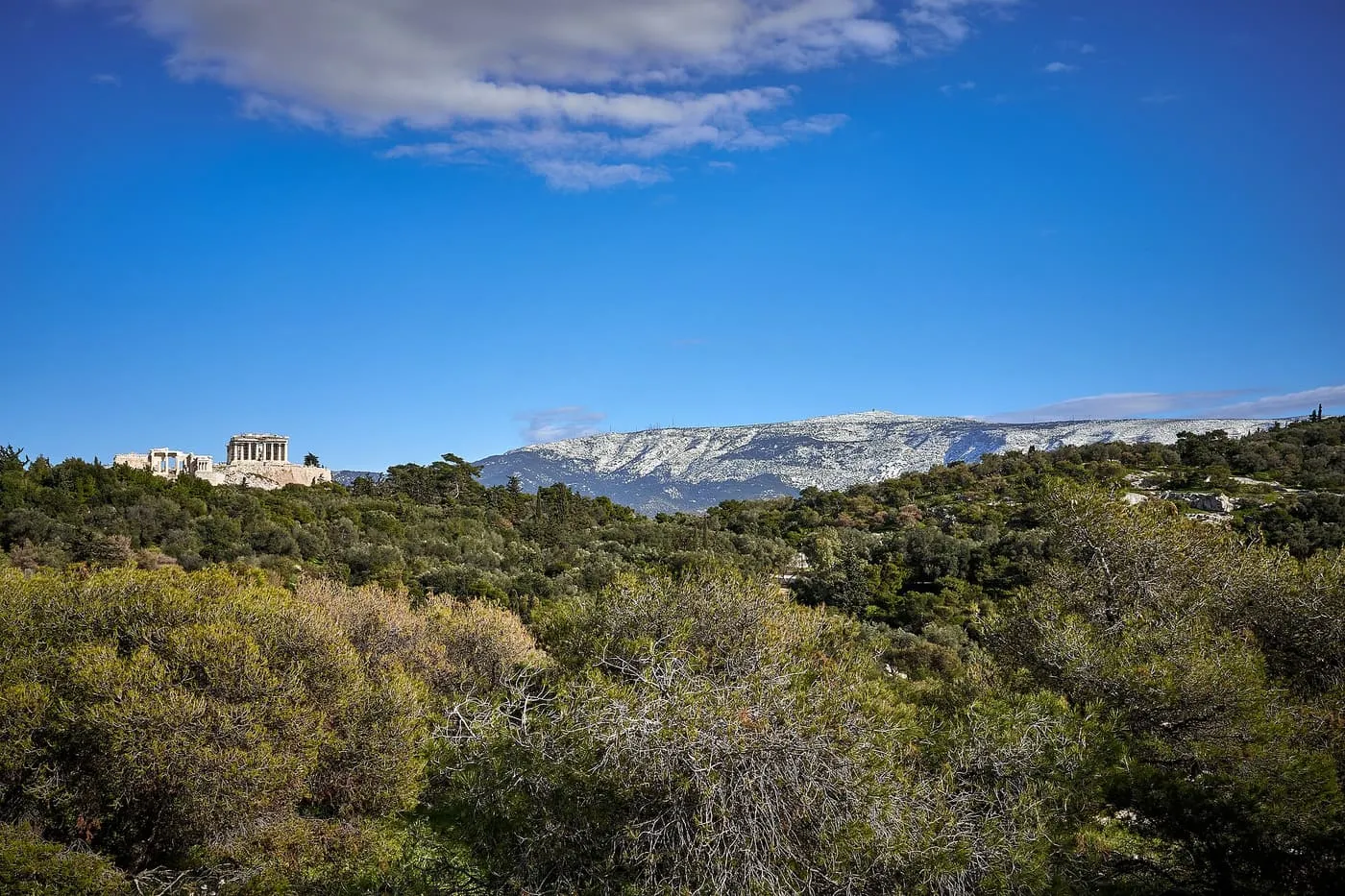 The Acropolis small on its hill at the left of a green wooded landscape, with the long ridge of Mount Hymettus dusted white with snow under a deep blue sky