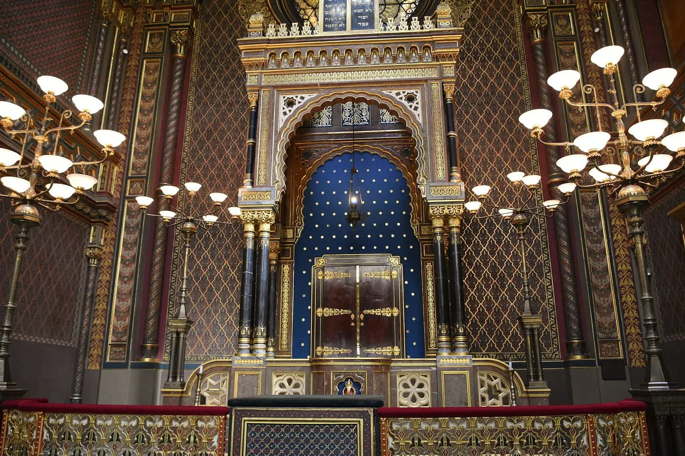 The interior of the Spanish Synagogue, with the gilded and blue Torah ark under a rose window, hanging chandeliers and carved galleries