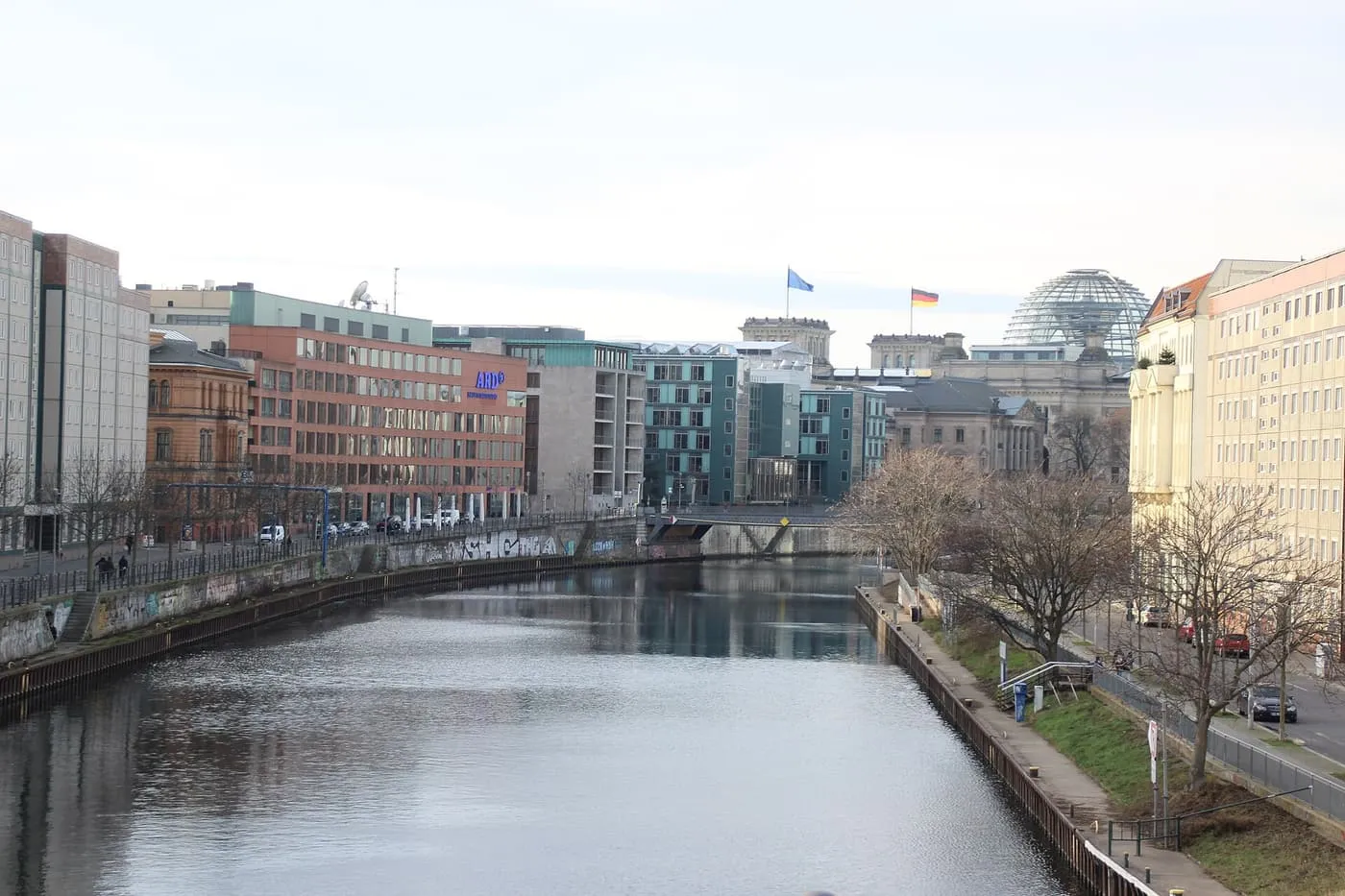 The Spree running between embankments with the glass Reichstag dome and two flags beyond a low bridge