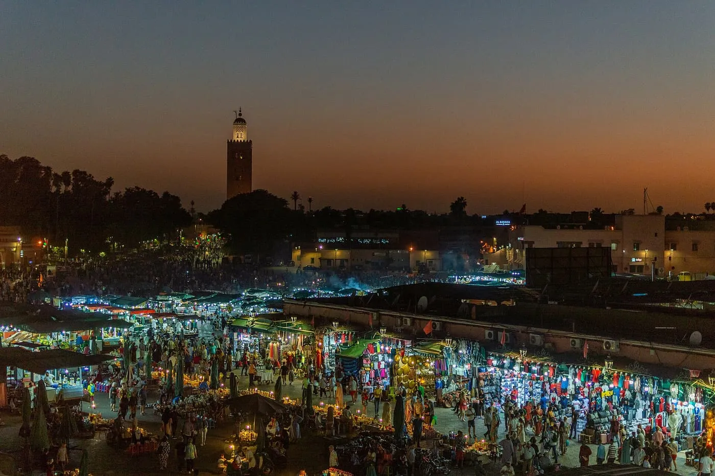 Jemaa el-Fnaa at dusk seen from above, with the lit food stalls and crowds below and the Koutoubia minaret on the dark horizon