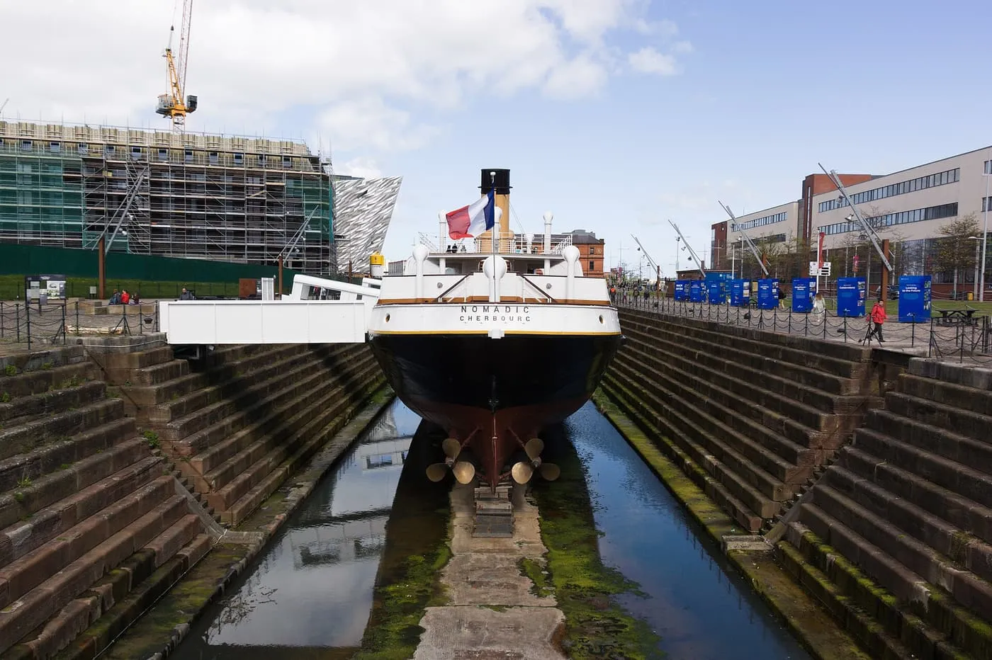 The stern of SS Nomadic in a stepped stone dry dock, with a French flag, and Titanic Belfast visible behind